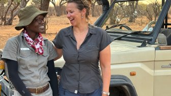 Zwei Frauen stehen lächelnd neben einem Jeep in der afrikanischen Savanne, umgeben von trockenen Bäumen und einem warmen Licht.