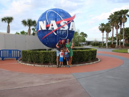 Eine Familie posiert fröhlich vor dem NASA-Symbol, umgeben von Palmen und einem gepflegten Garten.