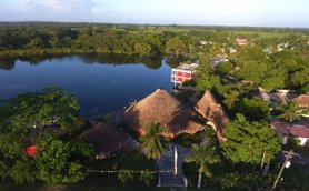 Eine Luftaufnahme zeigt das Lamanai Landings Hotel mit strohgedeckten Hütten am ruhigen Wasser eines Sees.