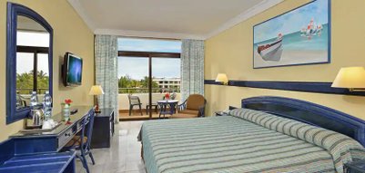 Ein helles Hotelzimmer mit einem großen Bett, einem Schreibtisch und einem Balkon mit Blick auf die tropische Landschaft.