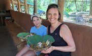 Eine Frau und ein Mädchen sitzen in einer rustikalen Umgebung und halten große Blätter mit buntem Essen in den Händen.