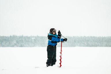 Ein Kind in blauer Winterkleidung bohrt mit einer roten Eisbohrmaschine in eine schneebedeckte Landschaft.
