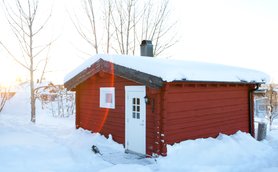 Eine rote Holzhütte steht in einer verschneiten Landschaft, umgeben von kahlen Bäumen und sanften Hügeln.