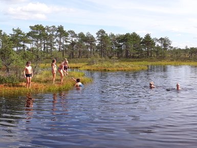 Sechs Kinder und Erwachsene genießen einen sonnigen Tag am Wasser, einige schwimmen, andere stehen am Ufer.
