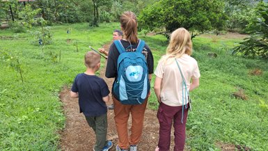 Eine Gruppe von Kindern und einem Erwachsenen geht auf einem Pfad durch eine grüne Landschaft voller Pflanzen und Bäume.