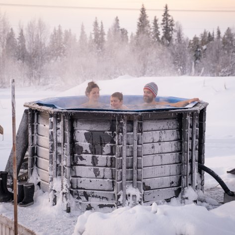 Drei Personen entspannen in einem heißen Whirlpool, umgeben von schneebedeckter Landschaft und frostigen Bäumen.