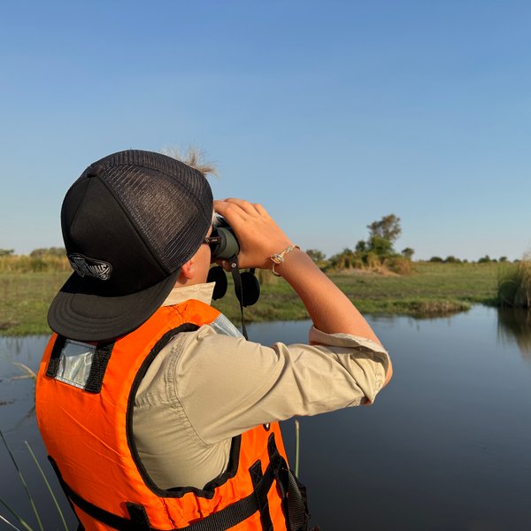 Ein Junge in einer orangefarbenen Weste beobachtet mit einem Fernglas die Landschaft am Wasser, umgeben von üppigem Grün.