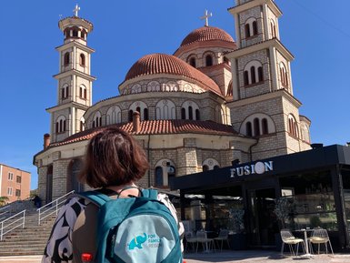 Eine Person mit einem Rucksack steht vor einer beeindruckenden Kirche mit mehreren Kuppeln und einem klaren blauen Himmel.