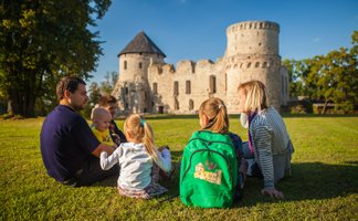 Eine Familie sitzt auf dem Gras vor einer alten Burg, während sie sich unterhält und die Sonne genießt.