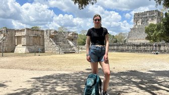 Eine junge Frau steht mit einer Tasche vor den beeindruckenden Ruinen von Chichen Itza, umgeben von grünem Laub und blauen Himmel.