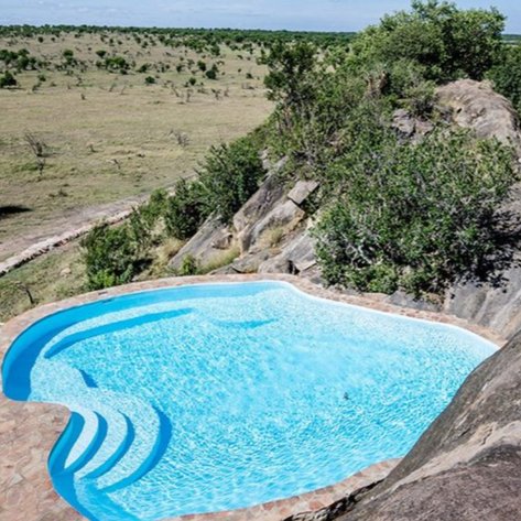 Ein glitzernder Pool schmiegt sich an einen Felsen, umgeben von üppigem Grün und der weiten Savanne im Hintergrund.