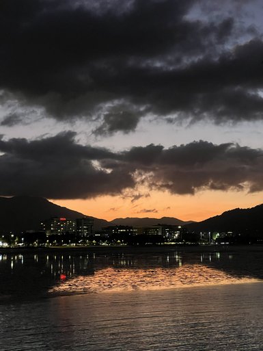 Dunkle Wolken über einem ruhigen Gewässer, mit orangefarbenem Sonnenuntergang, der die Silhouetten der Gebäude spiegelt.