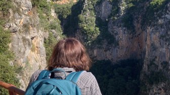 Eine Person mit einem blauen Rucksack steht an einem Geländer und blickt in eine tiefe Schlucht, umgeben von Bergen.