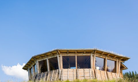 Ein modernes, rundes Holzgebäude mit großen Fenstern steht auf einer Wiese unter einem strahlend blauen Himmel.