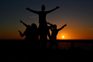 Fünf Silhouetten von Menschen, die fröhlich vor einem Sonnenuntergang posieren, während die Sonne am Horizont untergeht.
