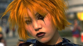 Eine Person mit leuchtend orangefarbenem Haar und dramatischem Make-up blickt ernst in die Kamera, umgeben von einer Menschenmenge.
