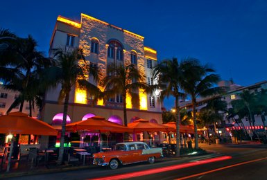 Ein historisches Hotel in Miami, beleuchtet von warmen Lichtern, umgeben von Palmen und orangefarbenen Sonnenschirmen.