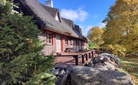 Ein traditionelles Reetdachhaus mit einer Holzveranda, umgeben von buntem Herbstlaub und einem blauen Himmel.