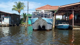 Zwei Boote liegen im Wasser vor einem angemalten Holzhaus mit Palmen im Hintergrund und strahlend blauem Himmel.