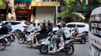 Eine belebte Straße in Hanoi, auf der viele Motorräder und Autos fahren, umgeben von grünen Bäumen und bunten Gebäuden.
