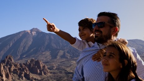 Eine Familie steht vor einer beeindruckenden Berglandschaft und zeigt auf etwas im Hintergrund, während sie lächeln.