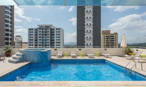 Ein glitzernder Pool auf einer Dachterrasse, umgeben von modernen Gebäuden und einem klaren blauen Himmel.