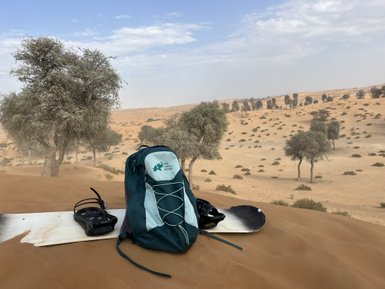 Ein Rucksack und ein Snowboard liegen auf einer Sanddüne, umgeben von vereinzelten Bäumen in einer Wüstenlandschaft.