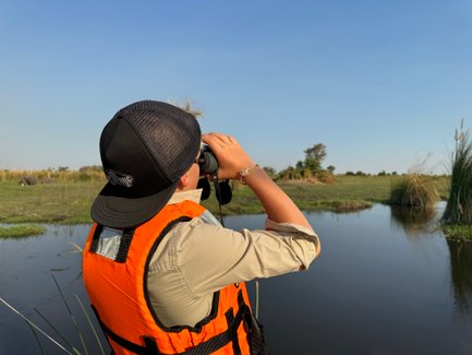 Ein Junge in einer orangefarbenen Weste beobachtet mit einem Fernglas die Landschaft am Wasser, umgeben von üppigem Grün.