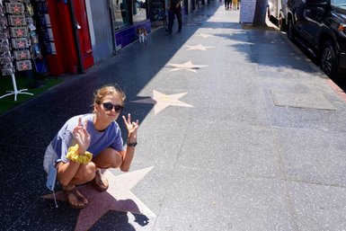 Eine junge Frau kniet auf dem Hollywood Boulevard und zeigt das Peace-Zeichen, umgeben von Sternen auf dem Gehweg.
