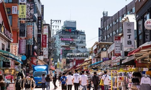 Eine belebte Einkaufsstraße in Hongdae, gesäumt von Geschäften und bunten Schildern, mit vielen Passanten im Sonnenlicht.