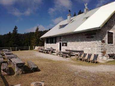 Eine gemütliche Berghütte aus Stein, umgeben von Bäumen, mit Holzstühlen und Tischen im Freien für Besucher.