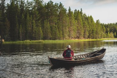 Ein Angler sitzt in einem Holzboot auf einem ruhigen See, umgeben von dichten, grünen Wäldern im Hintergrund.