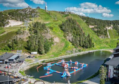 Ein lebhaftes Wasserpark-Set mit bunten Attraktionen auf einem klaren See, umgeben von grünen Hügeln und blauen Himmel.