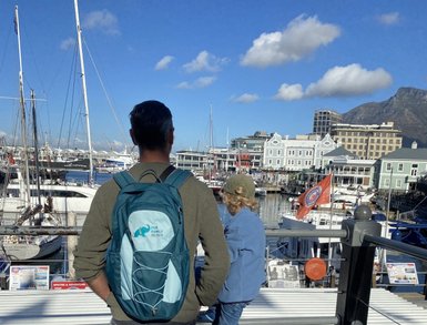 Ein Mann mit einem Rucksack steht am Hafen und blickt auf die Boote und die lebhafte Waterfront im Hintergrund.