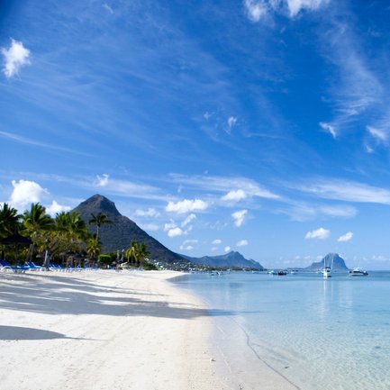 Ein ruhiger Strand mit feinem, weißem Sand und klarem Wasser, umgeben von Palmen und sanften Hügeln im Hintergrund.