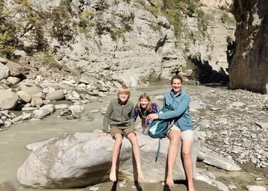 Drei Kinder und eine Frau sitzen auf einem großen Stein am Ufer eines schmalen Flusses, umgeben von steilen Felsen.