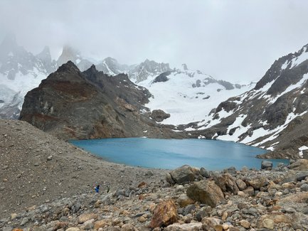 Ein klarer, blauer See liegt zwischen schroffen, schneebedeckten Bergen in einer nebligen Patagonien-Landschaft.