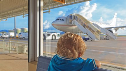 Ein kleines Kind mit blonden Locken sitzt am Fenster und beobachtet ein Flugzeug, das am Gate parkt.