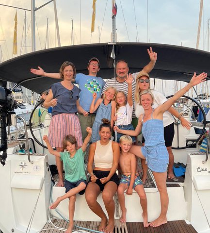 Eine fröhliche Gruppe von Erwachsenen und Kindern posiert auf einer Segelyacht, umgeben von Masten und einem schönen Himmel.