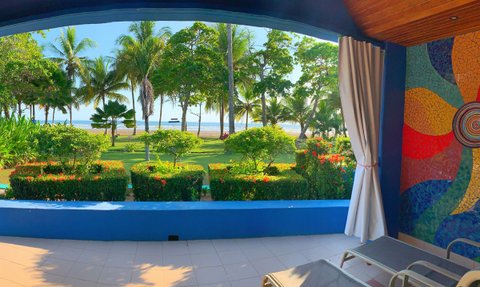 Ein Blick auf eine Terrasse mit bunten Pflanzen und Palmen, die zum Strand und zum Meer führen, unter klarem Himmel.