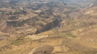 Eine Person sitzt auf einem Felsen und blickt über eine weite, trockene Landschaft mit sanften Hügeln und Feldern.
