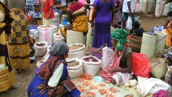 Ein belebter Markt mit Frauen, die um Waren wie getrockneten Fisch und Hülsenfrüchte handeln, umgeben von bunten Stoffen.