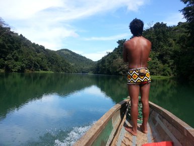 Ein Mann mit nacktem Oberkörper steht in einem Boot auf einem ruhigen Fluss, umgeben von üppigem Grün.