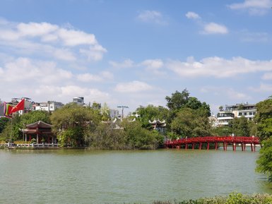 Eine rote Brücke erstreckt sich über den ruhigen Hoan-Kiem-See, umgeben von üppigem Grün und historischen Gebäuden.