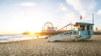 Ein Rettungsturm steht am Strand von Santa Monica, während die Sonne hinter dem Pier untergeht und die Wellen sanft an den Strand rollen.