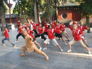 Eine Gruppe von Menschen in roten Jacken übt Kung Fu in einem traditionellen chinesischen Tempelbereich unter Bäumen.