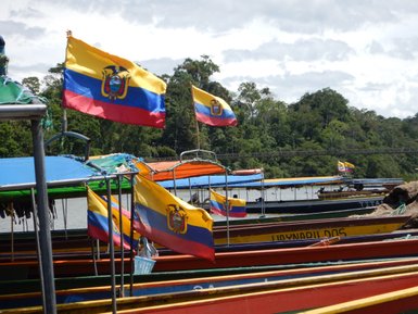 Bunte Boote mit ecuadorianischen Flaggen liegen am Ufer eines ruhigen Gewässers, umgeben von üppiger Vegetation.