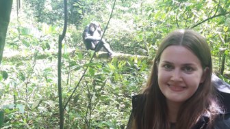 Eine Frau steht im Kibale Nationalpark, während ein Schimpanse im Hintergrund entspannt auf einem Ast sitzt.