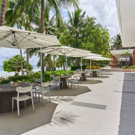 Ein schickes Restaurant am Strand, umgeben von Palmen und Sonnenschirmen, auf einem hellen Terrassenboden.