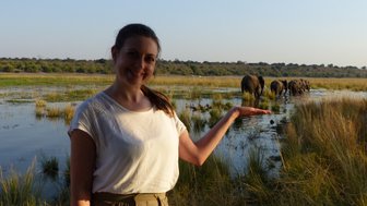 Eine lächelnde Frau steht am Ufer des Chobe-Flusses und zeigt auf eine Gruppe von Elefanten im Wasser.